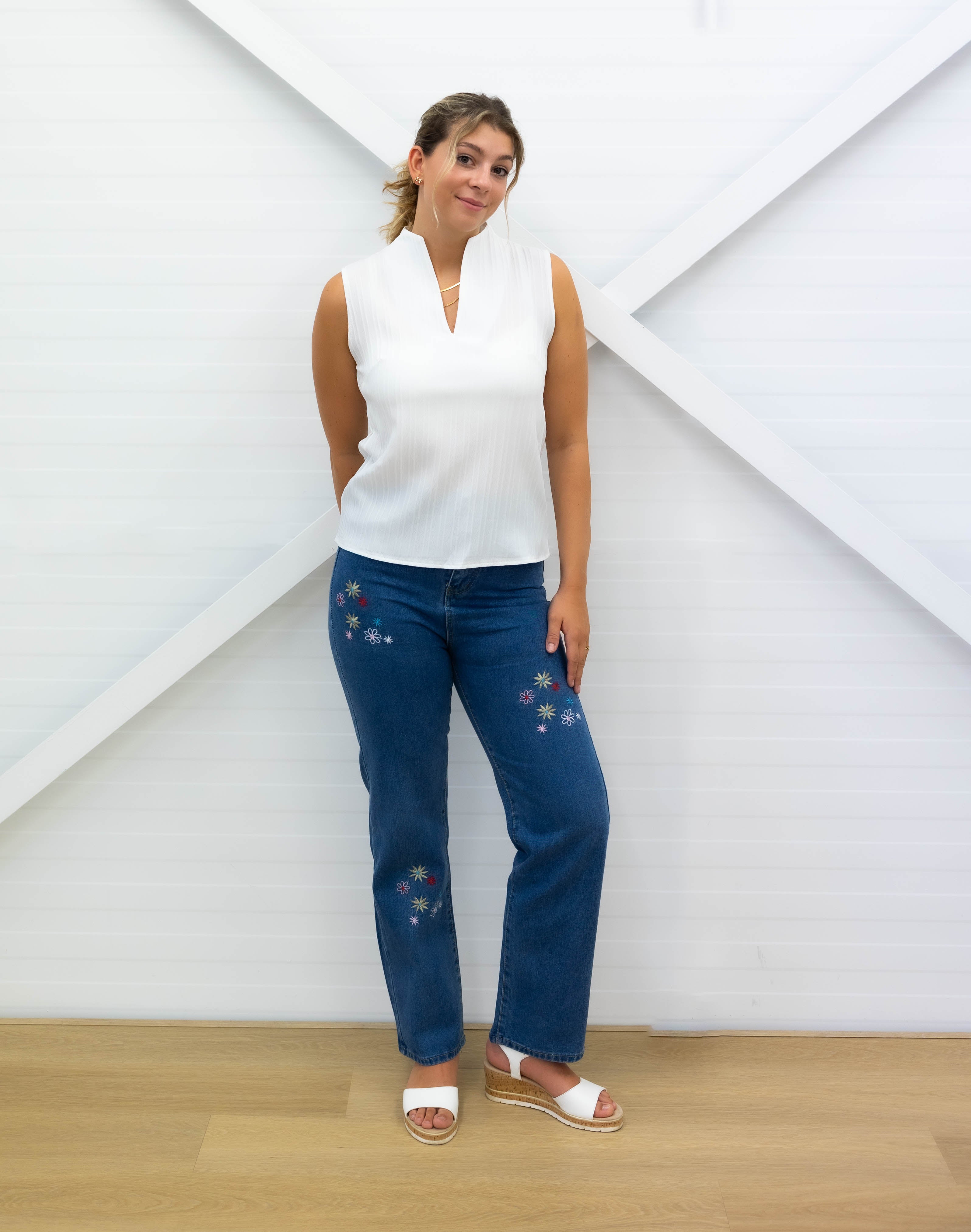 Woman wearing a white sleeveless top and blue jeans with floral embroidery against a white wall.